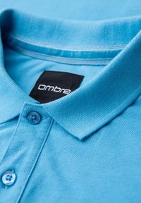 Ljusblå polo skjorta med en strukturerad tygliknande yta, med krage, två knappar och en svart etikett som står "ombre."