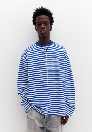 Bărbat tânăr cu păr scurt și creț, purtând o cămașă cu dungi albastre și albe cu mâneci lungi și blugi gri deschis, stând în picioare pe un fundal alb.