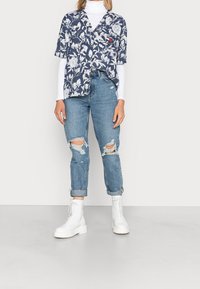 Camisa de manga corta de flores en tono marino con flores blancas, combinada con jeans claros desgastados. Botas tobilleras blancas con cordones completan el conjunto.