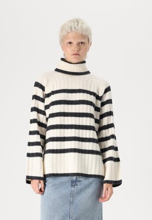 Streifenpullover in Creme und Schwarz, mit gerippter Textur, hohem Kragen und weiten Ärmeln. Kombiniert mit einem Jeansrock.