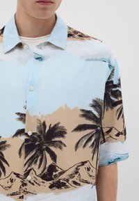 Camisa de manga corta hecha de tela ligera, con un fondo azul claro y un estampado de palmeras y montañas, botones y un collar relajado.