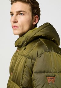 Oliwkowa kurtka puchowa z wysokim kołnierzem, zamek błyskawiczny i zapięcie na guziki oraz skórzaną naszywką z logo "Wrangler" na lewym rękawie. Gładka tekstura.