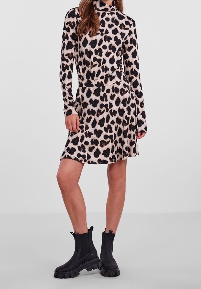 Kvinna iförd en långärmad klänning med leopardtryck, kombinerad med svarta kraftiga anklarstövlar, stående mot en enkel ljus bakgrund.