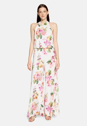 Mladá žena oblečená v biely overal s halter krkom s ružovo-zeleným kvetinovým vzorom, stojaca s rukami uvoľnenými pozdĺž tela.