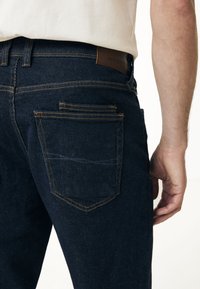 Mörkblå denimjeans med en synlig läderetikett vid midjan och kontrasterande orange sömmar på bakfickorna, med en slät yta.