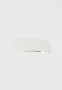 Semelle de sneaker blanche présentant un motif de grip texturé, une forme arrondie, une finition lisse et une inscription discrète de la marque au centre.