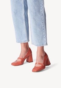 Chaussures à talons hauts en cuir verni corail, avec un bout carré et un talon épais, présentant une sangle sur le coup de pied et un petit accent doré.
