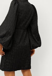Schwarzes Kleid aus strukturiertem Stoff, mit Ballonärmeln, tailliertem Schnitt und solidem Saum. Das Design weist subtile Variationen im Stoffmuster auf.
