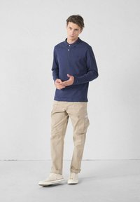 Polo a maniche lunghe blu navy con colletto e dettaglio del logo, abbinato a pantaloni cargo beige con più tasche e orli arrotolati.