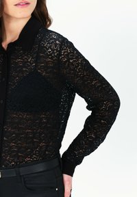 Camicia nera di pizzo con maniche lunghe e colletto. Presenta un motivo floreale e chiusura con bottoni, abbinata a un tessuto nero liscio sottostante.