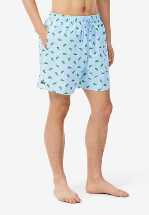 Man draagt lichtblauwe zwemshorts met kleine groene krokodillenpatronen, elastische taille, trekkoord en zakken, staat blootsvoets op een witte achtergrond.
