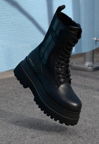 Stivali piattaforma neri in pelle lucida con punta rotonda, chiusura con lacci e logo sul lato. Suola in gomma testurizzata.