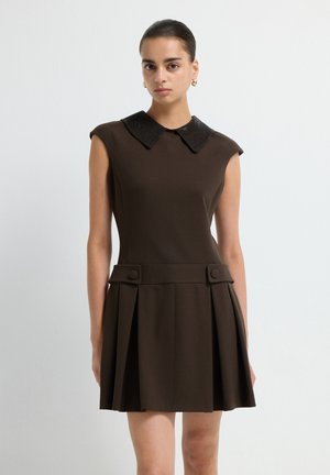Frau in einem ärmellosen dunkelbraunen Kleid mit einem schwarzen strukturierten Kragen, plissiertem Rock und knopfseitigem Detail, stehend vor einer schlichten Wand.