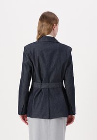 Blazer din denim închis, croit pe măsură, cu curea în talie, umeri structurați și o textură fină, stilizat cu un guler la gât și ventilație pe spate.
