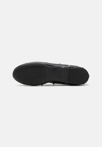 Zapato plano negro con suela de goma texturizada, que presenta acentos de tachuelas metálicas a lo largo de los lados. Parte superior de cuero liso y diseño minimalista.