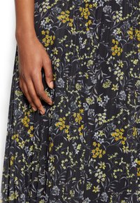 Floral dress with a black background, featuring yellow and light blue flowers. Hand gently grasps the fabric, showcasing its pleated texture.