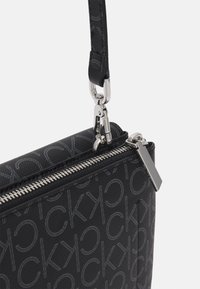 Černá kabelka s reliéfním vzorem loga, stříbrnými detaily, zipovým uzávěrem a texturovaným povrchem. Obsahuje odnímatelný popruh pro všestranné nošení.
