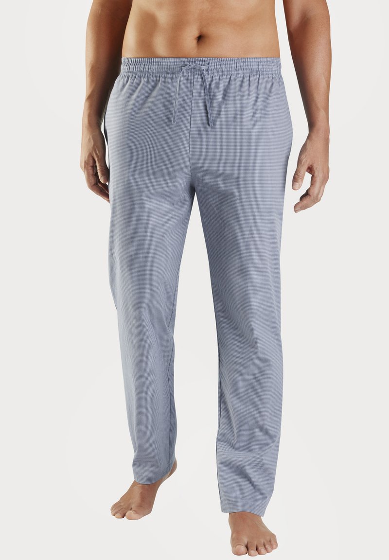 Muž oblečený v svetlomodrých károvaných pyžamových nohaviciach s obvodovým pásom, stojaci bosý proti bielej pozadí.