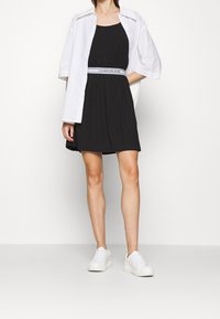 En modell bär en svart klänning med ett Calvin Klein-bälte och en vit skjorta, tillsammans med vita sneakers, poserande mot en enkel bakgrund.