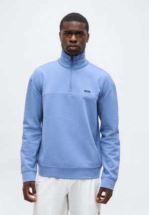 Muž nosi svetlomodrý pulóver s polovičným zipsom a logom "BOSS" na hrudi, v bielych nohaviciach, stojaci na jednofarebnom pozadí.