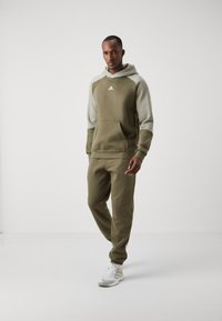 Felpa verde oliva con maniche grigio chiaro e tasca frontale, abbinata a pantaloni sportivi verde oliva e scarpe da ginnastica bianche.