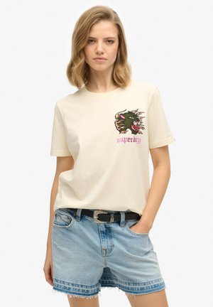 T-shirt en coton crème avec un graphique d'un dragon vert et rouge, associé à un short en denim bleu délavé et une ceinture noire.