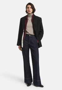 Frau, die in einem geschichteten Outfit steht, bestehend aus einem beigen Rollkragenpullover, einer burgunderroten Zip-Jacke, einem schwarzen Blazer, dunklen, weiten Jeans und schwarzen Schuhen.