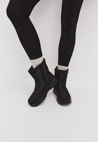 Botas negras con cremallera, con un acabado texturizado, detalles acolchados y suelas de goma gruesas. Combinadas con leggings negros y calcetas de color gris claro.