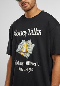 Černé bavlněné tričko s grafikou naskládaných stohů bankovek v hodnotě sto dolarů se žlutou stuhou. Text zní: "Peníze mluví mnoha různými jazyky" bílou barvou.