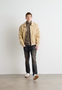 Chaqueta bomber beige sobre un cuello de tortuga gris, combinada con jeans oscuros y zapatillas blancas. El modelo se encuentra de pie contra un fondo blanco liso.