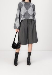 Nainen, joka wearing harmaan ja vaaleanharmaan rombikuviollisen neuleen, tummanharmaat laskostetut polvenpituiset shortsit, mustat saappaat ja pitää mustaa käsilaukkua.