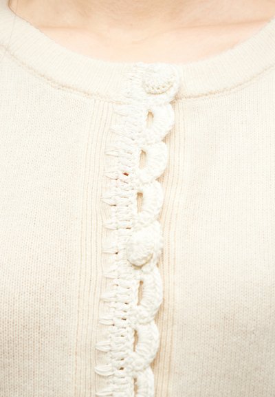 Κοντινό πλάνο κρεμ πλεκτού ζακέτας με δαντελένιο διακοσμητικό κρόσσι και κουμπιά καλυμμένα με ύφασμα κατά μήκος του κεντρικού μπροστινού ντεκολτέ.
