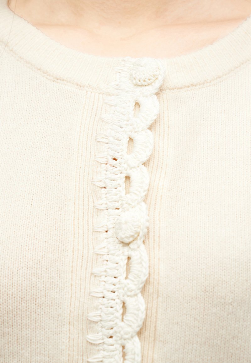 Close-up van een crème gebreide cardigan met gegolfde haakrand en met stof beklede knopen langs de halslijn aan de voorkant.