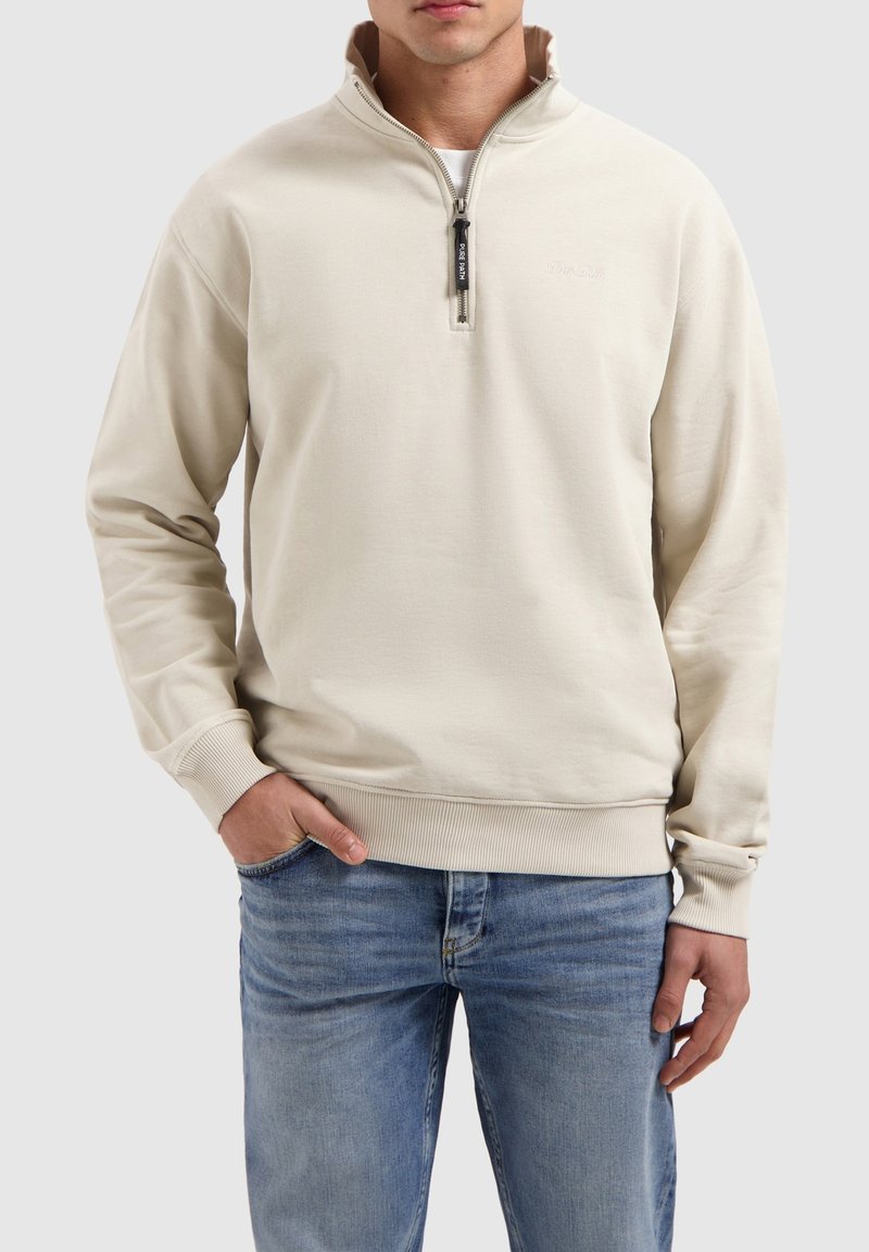 Beige sweatshirt met een kwart-ritssluiting, geribbelde mouwen en onderkant, met een klein logo op de borst. Draag over lichtblauwe jeans.