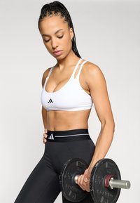 Balta sportinė liemenėlė su plonomis petnešėlėmis, juodas logotipas; derinama su aukštos liemens juodomis tamprėmis su balta akcentuota juostele; laiko hantelį.