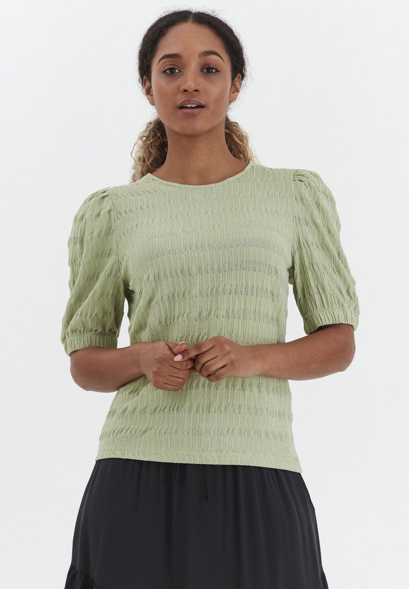 Blusa verde chiaro con texture, maniche corte a palloncino e scollo rotondo, realizzata in tessuto stropicciato. Finitura liscia e vestibilità rilassata.