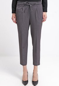 Pantalon gris ajusté et effilé avec une taille à cordon, présentant des plis et une texture lisse, associé à des chaussures à talons noires.