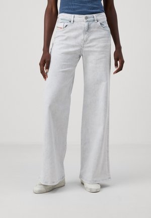 Person iført lyseblå vidde jeans og hvide sneakers, stående mod en ensfarvet lys grå baggrund.