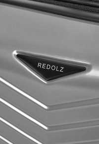 Silberner Hartschalenkoffer mit Chevron-Muster, einem schwarzen dreieckigen Logo mit "REDOLZ" und einem Reißverschlussfach.
