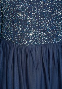 Námořnické modré šaty s korzetem pokrytým flitry, který obsahuje lesklé stříbrné a modré disky, a sukni z plisovaného tylu pro texturu a objem.