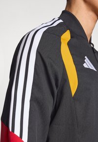 Svart treningsjakke med hvite og gule striper på skuldrene, med en rød detalj på underarmen og glidelås foran.