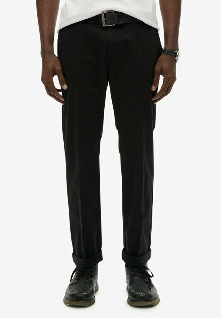 Pantalones negros hechos de una tela resistente, con un corte recto. Llevados con un cinturón negro y zapatos negros. Los puños enrollados ligeramente en los tobillos.