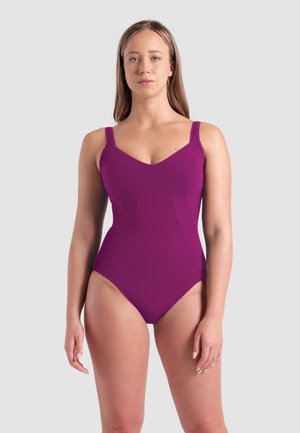 Donna con i capelli lunghi che indossa un costume intero viola, in piedi davanti a uno sfondo grigio chiaro uniforme.