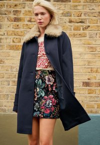 Tmavomodrý vlnený kabát s umelým kožušinovým golierom, nasadený na krémovom topu s červeným písmom a sukni s kvetinovým vzorom v tmavých a výrazných farbách.