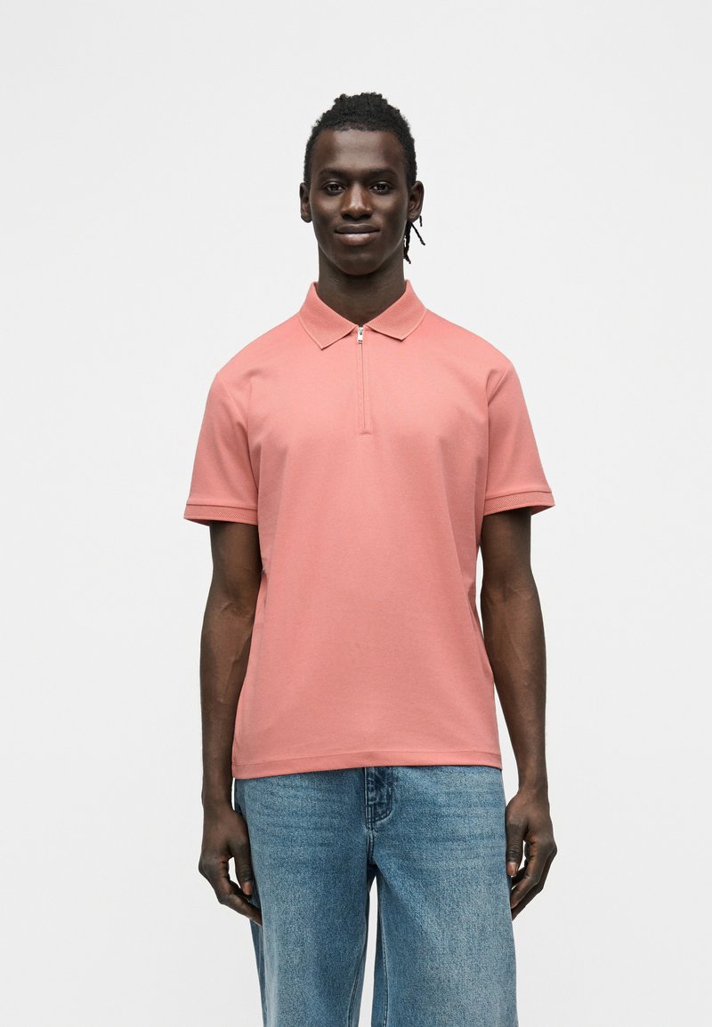 Άνδρας στέκεται προς τα εμπρός, φορώντας κοντομάνικο ροζ πολο πουκάμισο με φερμουάρ και ανοιχτό μπλε τζιν, μπροστά σε απλό λευκό φόντο.