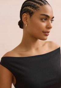 Top negro sin hombros con una textura suave. La modelo tiene el cabello trenzado y lleva un pequeño aro de oro. Fondo suave.