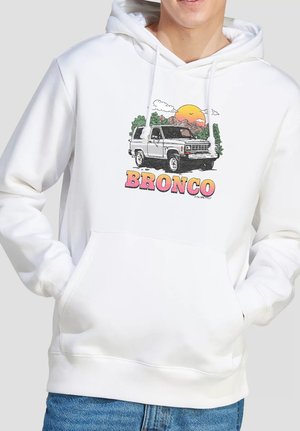 Person trägt einen weißen Hoodie mit einem Motiv eines klassischen Bronco-SUV, Sonnenuntergang, Bäumen und dem Wort "BRONCO" in pinken und orangen Buchstaben.