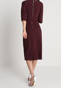 Robe bordeaux ajustée avec manches trois-quarts, fermeture éclair au dos, ceinture à la taille ; tissu lisse et longueur au genou.