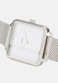 Vierkante zilveren horloge met een witte wijzerplaat, zilveren uurmarkeringen en een mesh band. Heeft een kleine kroon aan de zijkant voor aanpassingen.