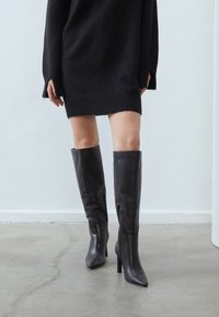 Bottes en cuir noir montant jusqu'au genou, avec des pointes et des talons aiguilles, dotées d'une fermeture zippée sur le côté et d'une texture lisse.
